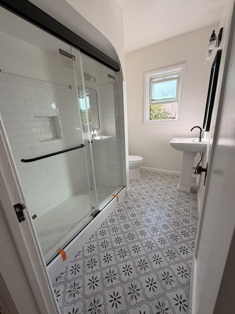 Completed Bandini restroom remodel with a white subway tile shower, black framed glass enclosure, pedestal sink and patterned floor tile